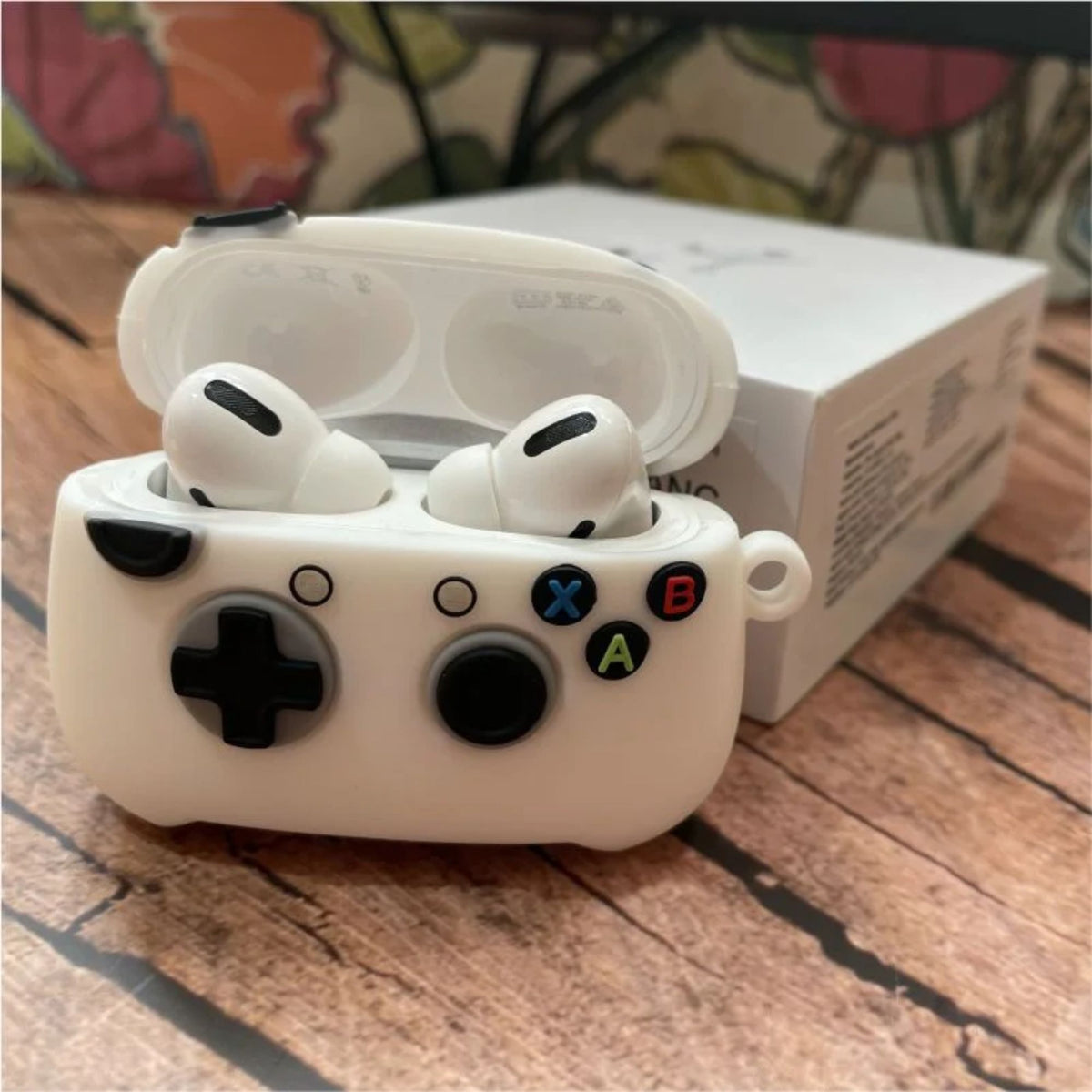 A white silicone case designed to look like a retro game controller, shown with a pair of white wireless earbuds inside its open charging case. The case features black controls and colored A, B, X, and O buttons.