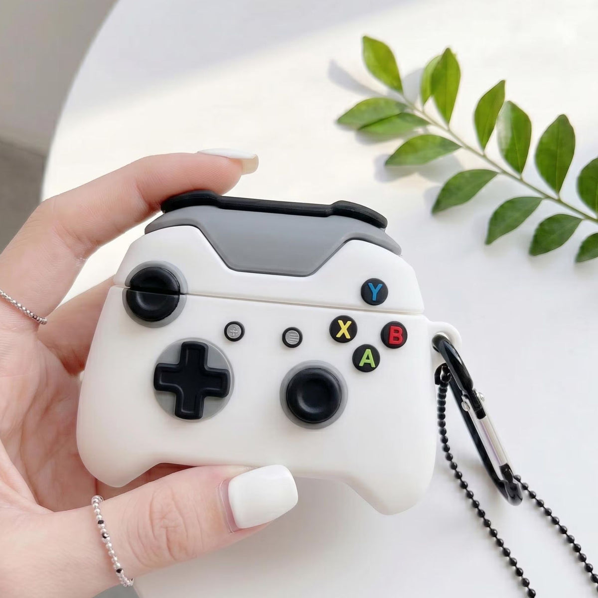 A hand holds a white and gray game controller-style case with black controls and a black ball chain strap, photographed on a white surface with a green leaf in the background.